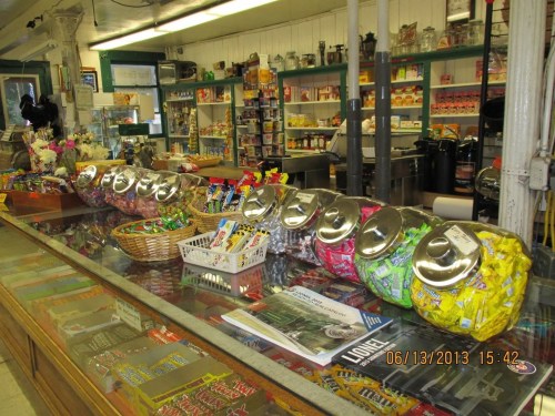 A vintage store interior featuring a glass display case with colorful candy jars and shelves stocked with various goods.