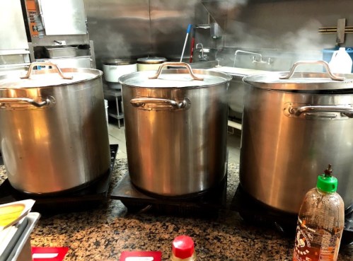 Three large pots on a stove, steaming, in a commercial kitchen setting. Bottles of sauce are visible in the foreground.