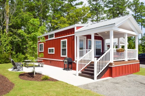 A red tiny house with white trim, surrounded by trees, featuring a porch, outdoor seating, and a grill.