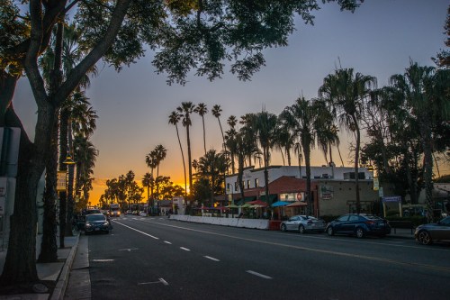 Sunset over a palm-lined street with cars parked along the side and buildings in the background.