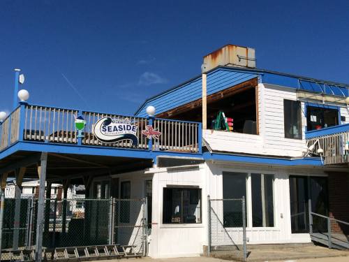 A seaside building with a blue roof, featuring a sign and outdoor seating, under a clear blue sky.