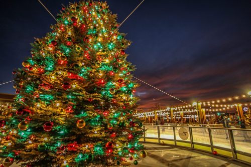 A brightly lit Christmas tree adorned with colorful ornaments, set against a twilight sky and an ice skating rink.