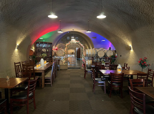 Interior of a cozy restaurant with wooden tables, chairs, and wine barrels, featuring colorful lighting and a warm atmosphere.