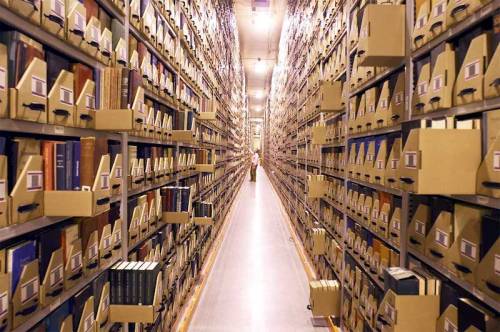 A long aisle between tall shelves filled with boxes and books in a large archive or library.