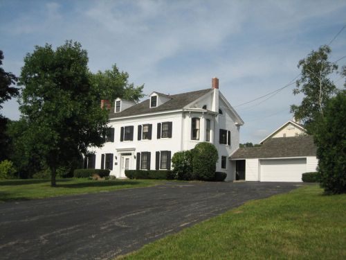 A white two-story house with black shutters, surrounded by greenery and a paved driveway leading to a garage.