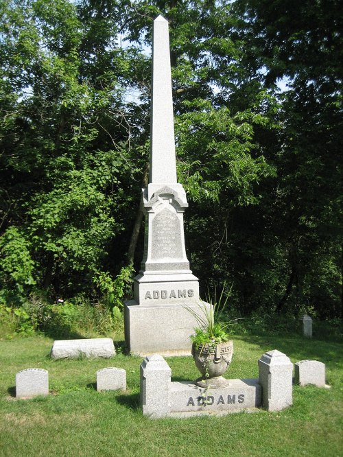 A tall granite obelisk monument with the name "ADDAMS" surrounded by smaller gravestones in a green, wooded area.