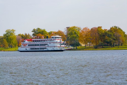 A large riverboat docked near a tree-lined shore with autumn foliage, reflecting in the calm water.