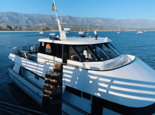 A white yacht docked by the shore, with mountains in the background and boats in the water.