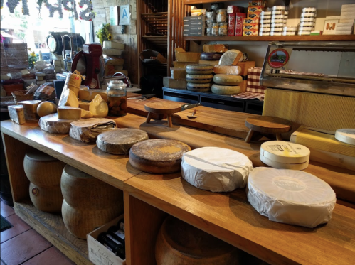 A wooden table displays various types of cheese, with shelves of cheese wheels and jars in the background.