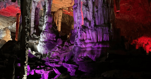 Colorful stalactites and stalagmites illuminated in a cave, showcasing vibrant purple and red lighting against rocky formations.