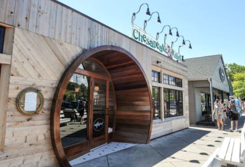 Exterior of a wooden restaurant with a circular entrance, featuring a sign that reads "Chesapeake Grill." Sunny day.