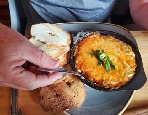 A hand holds a fork near a sizzling dish of melted cheese, accompanied by slices of toasted bread on a plate.