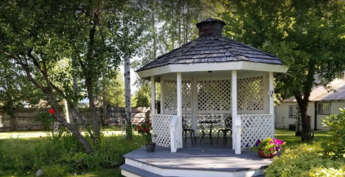 A white gazebo with a shingled roof surrounded by greenery and flowers in a sunny garden setting.