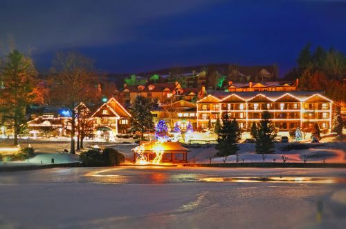 A winter scene featuring a cozy lodge illuminated with holiday lights, surrounded by snow and trees at dusk.