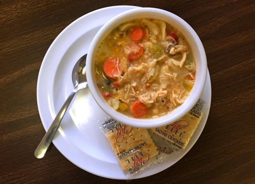 A bowl of chicken noodle soup with vegetables, served on a plate with a spoon and a packet of saltine crackers.