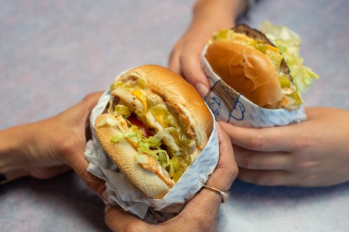 Two hands hold burgers wrapped in paper, one with lettuce, tomato, and pickles, the other with a plain bun.