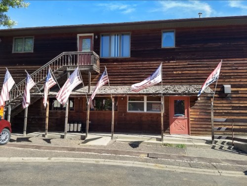 A wooden building with a porch, featuring multiple American flags and a red door, set against a clear blue sky.