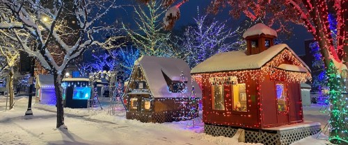 Colorful holiday lights adorn snow-covered houses in a winter scene, with trees illuminated in the background.