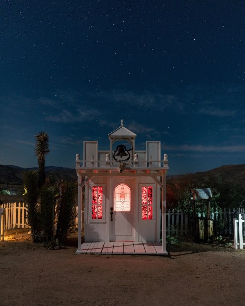 A small, white building with red-lit windows under a starry night sky, surrounded by desert landscape and a fence.