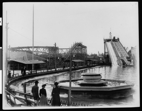 A vintage amusement park scene featuring a roller coaster, water slide, and a fountain, with people in period clothing.