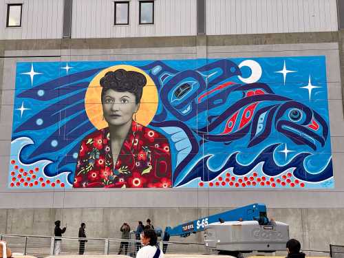 A vibrant mural featuring a woman with floral patterns, surrounded by blue waves and stylized fish, set against a gray wall.