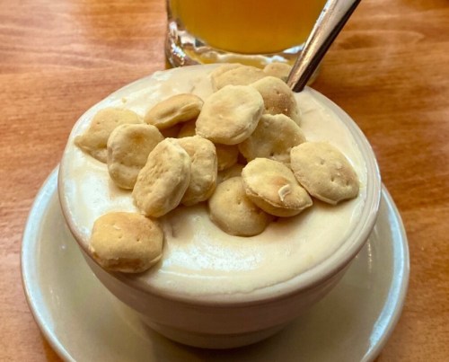 A small bowl of creamy dessert topped with crunchy snacks, placed on a wooden table with a drink in the background.