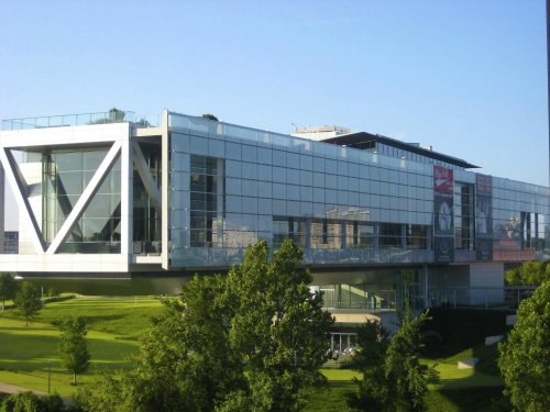 Modern glass building with a green landscape, featuring large windows and a balcony, under a clear blue sky.