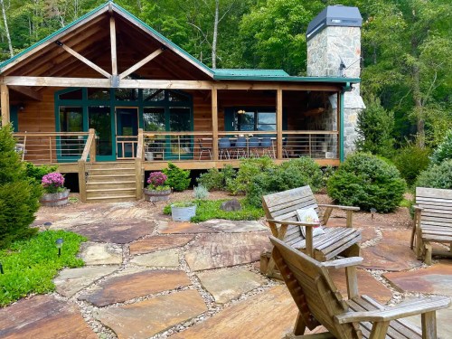 A rustic wooden cabin surrounded by greenery, featuring a stone chimney and a patio with wooden chairs.