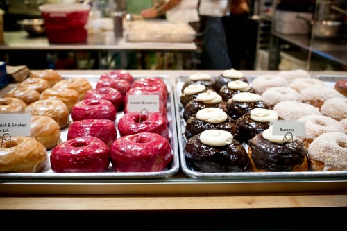 A display of assorted donuts, including glazed, lemon blueberry, chocolate, and buckeye varieties, in a bakery setting.