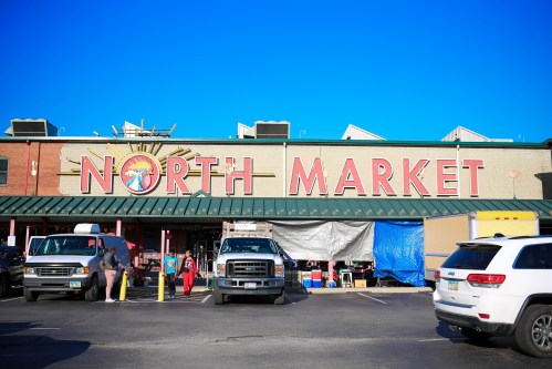 A vibrant market building with "NORTH MARKET" signage, surrounded by parked vehicles and clear blue skies.