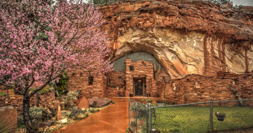 A stone structure built into a rocky cliff, surrounded by blooming pink trees and a fenced garden area.