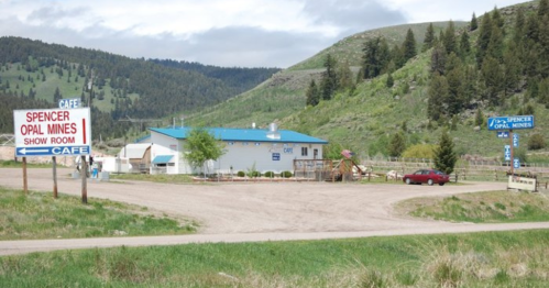 A small building with a blue roof, labeled "Cafe," surrounded by green hills and trees, near a dirt road.