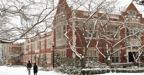 Two people walk through a snowy landscape near a brick building with trees and lampposts in winter.