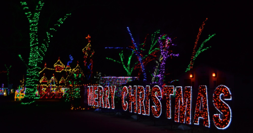 Colorful Christmas lights illuminate trees and decorations, with a large "MERRY CHRISTMAS" sign in the foreground.