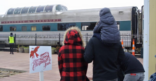 A family watches a train at a station, with a sign for a holiday event in the foreground.