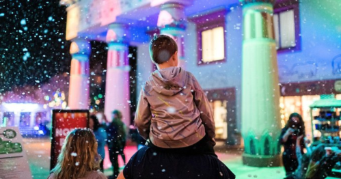A child on an adult's shoulders watches colorful lights and falling snow in a festive nighttime setting.