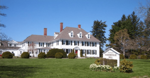 A large white colonial-style house with a manicured lawn and trees, featuring a sign for "Woodstock Hill."