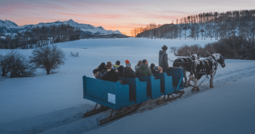 A horse-drawn sleigh carries passengers through a snowy landscape at sunset, with mountains in the background.