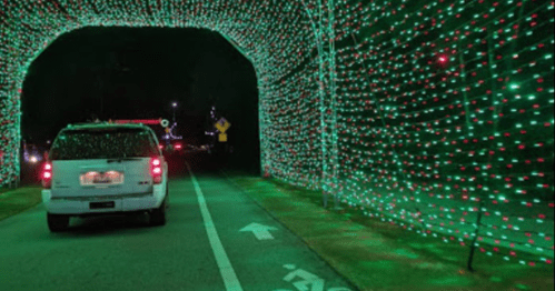 A car drives through a tunnel of colorful holiday lights at night, with green and red lights illuminating the scene.