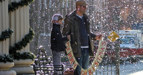 A man and a child stand in falling snow, holding a "Merry Christmas" banner in a festive outdoor setting.