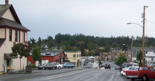 A quiet street in a small town, lined with parked cars and buildings, under a cloudy sky. Trees are visible in the background.