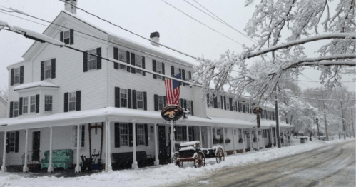 A historic white building covered in snow, with an American flag and a vintage wagon in front, on a quiet street.
