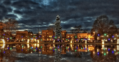 A festive scene with a lit Christmas tree reflecting in water, surrounded by colorful lights and a cloudy evening sky.