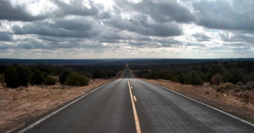 A long, empty road stretches into the distance under a cloudy sky, surrounded by sparse vegetation and hills.
