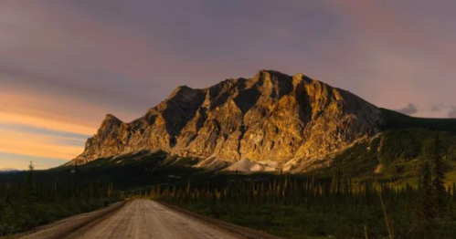 A dirt road leads towards a majestic mountain illuminated by a colorful sunset sky.