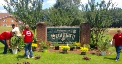 People planting flowers and shrubs around a "Welcome to Seminary" sign in a green, sunny area.