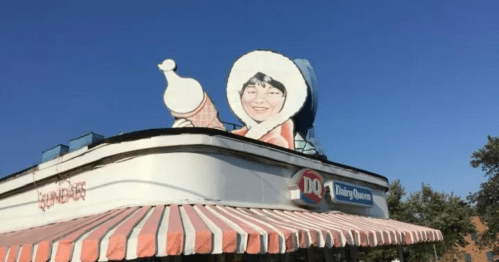 A Dairy Queen building with a large, smiling ice cream mascot on the roof against a clear blue sky.
