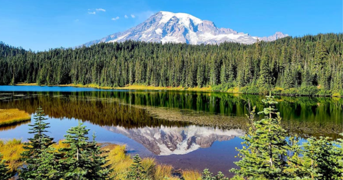 A serene lake reflects a snow-capped mountain surrounded by lush green forests under a clear blue sky.