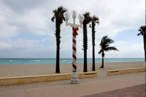 A beachside scene featuring a striped lamp post with a bow, surrounded by palm trees and a cloudy sky.