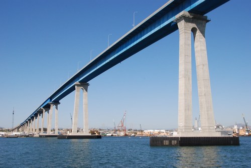 A blue bridge spans over water, supported by tall concrete pillars, with a clear blue sky in the background.